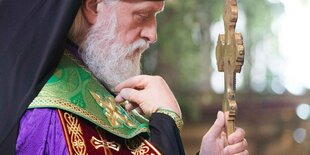 Ein Mann mit schwarzer Haube und religiöser Kleidugn hält ein goldenes Kreuz