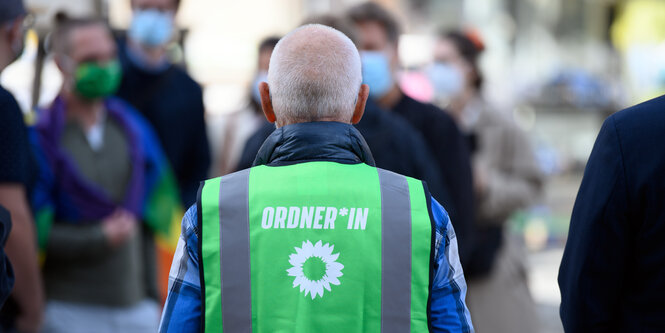 Eine Person mit grauen Haaren von hinten trägt eine grüne Weste mit weißer Aufschrift «Ordner*in».