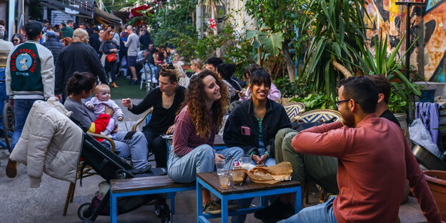 Menschen sitzen in einem Straßencafé mitten in einer vollen Straße in Tel Aviv, sie lachen und trinken Kaffee.