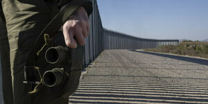 Ein Soldat hält ein Fernglas vor einem Grenzzaun
