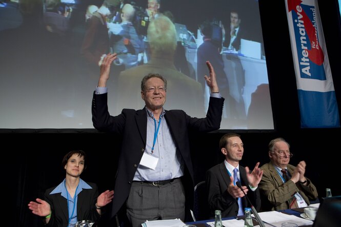 Frauke Petra, Konrad Adam, Bernd Lucke und Alexander Gauland sitzen vor einer Videowand an einem Tisch. Konrad Adam steht und grüßt mit geöffneten Armen