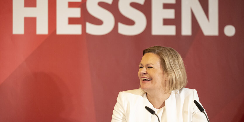 Nancy Faeser hat mittellange blonde Haare, trägt ein weißes Sakko, lacht und schaut zur Seite