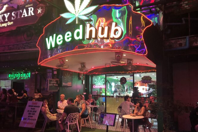 Touristen sitzen am ABend in einer Kneipe auf der Straße, Weedhub heißt der Laden mit Cannabisblatt in Leuchtschrift