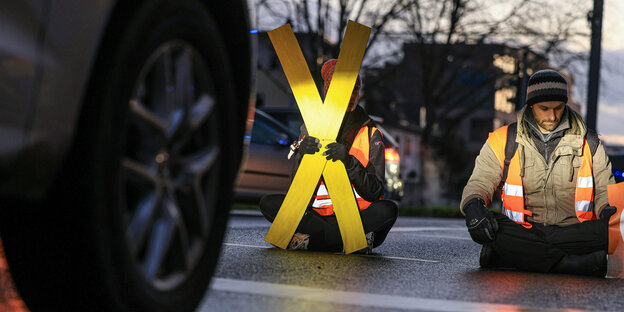 Zwei Klimaaktivisten sitzen auf der Straße vor einem Auto, eine Person hält ein großes X in der Hand