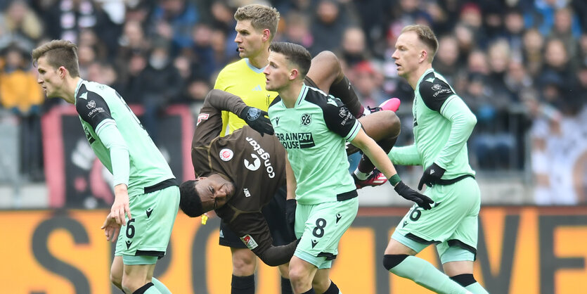 Ein Fußballspieler im braunen Trikot liegt quer in der Luft zwischen anderen Spielern im grünen oder gelben Trikot