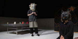 Zwei Schauspielende auf einer Bühne tragen einen Schafs- und einen Wolfskopf auf den eigenen Häuptern