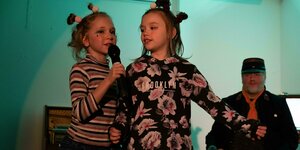 Zwei Mädchen mit kleinen Haardutts am Mikrofon, im Hintergrund ein erwchsener Darsteller