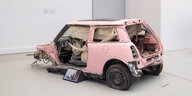 Ein zerstörter, rosafarbener Kleinwagen, offenbar nach einem Unfall. Davor ist ein Bildschirm platziert