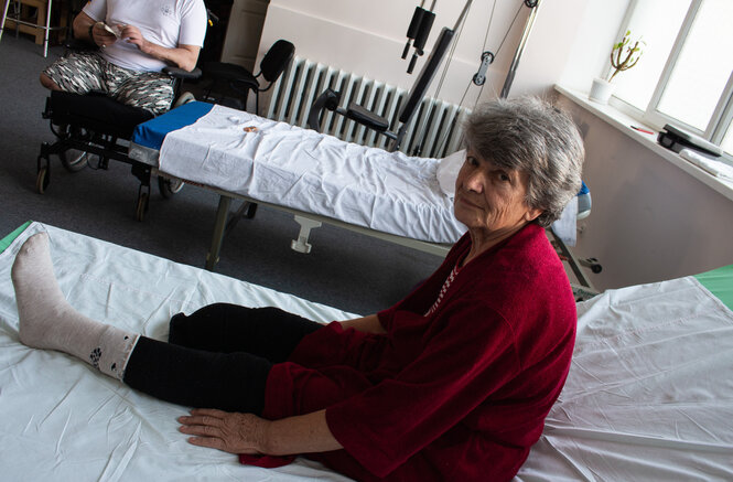 Eine Frau mit grauen Haaren sitzt auf einem Bett. Sie trägt einen roten Pullover und schwarze Leggins. Ihr rechtes Bein ist ab dem Knie amputiert