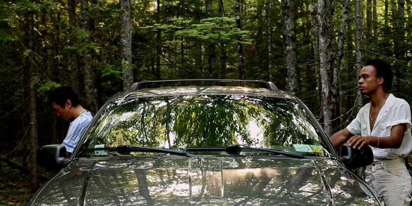 Filmszene: Ein Auto steht in einem Wald. Neben der Fahrer- und der Beifahrertür stehen rechts und links jeweils ein Mann in Sommerkleidung. Die Person links im Bild hat sich vom Auto abgewendet. Der Mann rechts im Bild hat die Hand auf den Autospiegel gelehnt und schaut wie in Gedanken ins Leere.