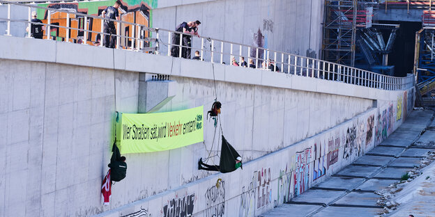 Zwei Klimaaktivisten seilen sich von einer Betonmauer auf der A100 Baustelle ab. Sie halten ein Transparent: "Wer Straßen sät, wird Verkehr ernten". Polizisten gucken.