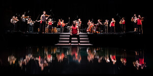 Orchestermusiker in schwarzen Hosen und roten shirts spiegeln sich auf einer dunklen Fläche