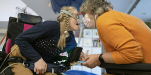 Ein Kind mit Beatmungsgerät hält die Hand einer grauhaarigen Frau. Beide sitzen in einem Rollstuhl.