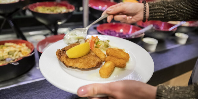 Im Vordergrund ein Teller mit Essen auf den gerade noch mehr Essen aufgegeben wird, im Hintergrund ein Buffet