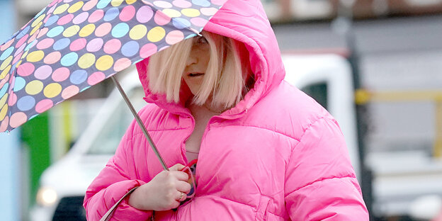 Eine Frau in pinker Jacke hält einen Regenschirm vor ihrem Gesicht