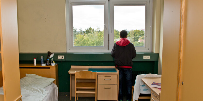 Ein Junge steht allein am Fenster eines Zimmers und blickt hinaus.