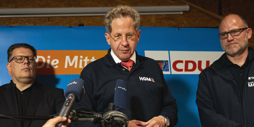 Hans-Georg Maaßen vor einem Mikrofon, auf seinen Pulli sind seine Initialien gestrickt