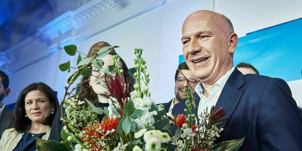 Kai Wegner steht lachend mit einem Blumenstrauß auf einer Bühne, umringt von Anhängern