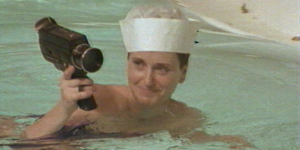Eine Frau mit Matrosenhut schwimmt in einem Pool und hält eine Super-8-Kamera in der Hand, die sie über der Wasseroberfläche in die Luft hält