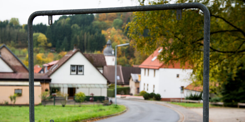 Durch den Rahmen eines Ortschildes, dessen Inhalt aber fehlt, sind unscharf Gebäude und eine Kirche zu sehen, im Hintergrund Berge