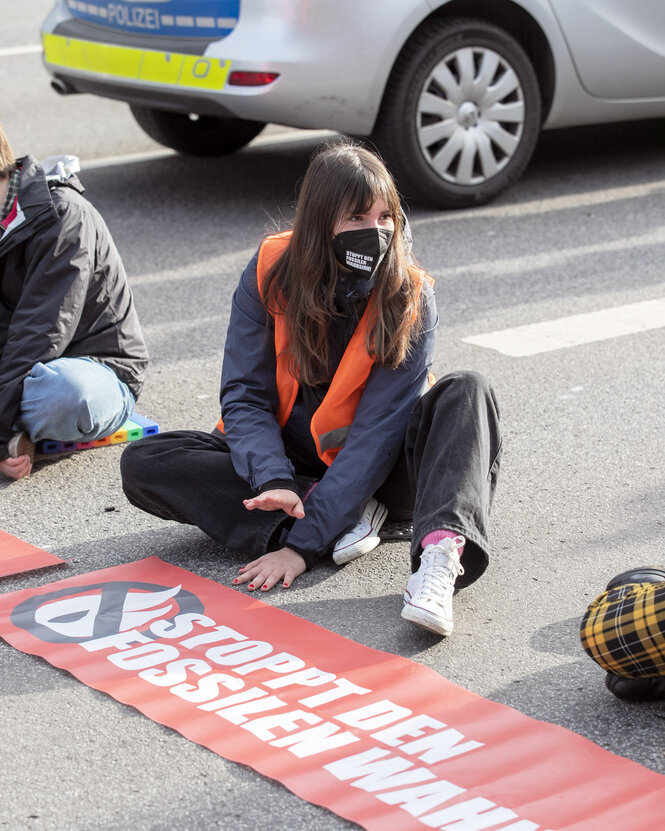 Hinrichs hockt vor einem Banner der Letzten Generation auf der Straße. Sie trägt einen schwarzen Mundschutz und eine orangefarbene Warnweste.