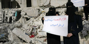 Zwei vollverschleierte Frauen stehen vor einem zerstörten Haus und halten ein Protestplakat gegen die UN in die Kamera