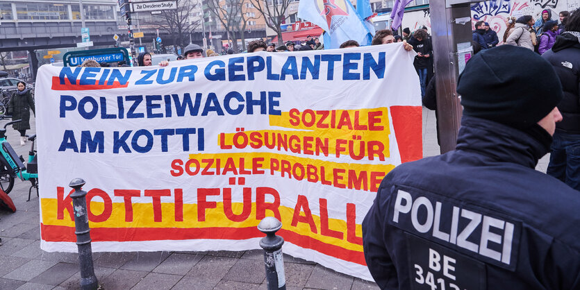 „Nein zur geplanten Polizeiwache am Kotti - soziale Lösungen für soziale Probleme - Kotti für Alle“ steht auf dem Transparent linker Gruppen, davor steht ein Polizist. Am Kottbusser Tor in Berlin-Kreuzberg wird an diesem Tag eine umstrittene neue Polizeiwache eröffnet.