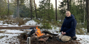 Ein Mann mit Glatze sitzt im Wald in Winterjacke vor einem brennenden Lagerfeuer im Schnee . Es ist taghell. Im Hintergrund sind vor allem Nadelbäume zu sehen.