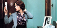 Eine braunhaarige Frau in Bluse und Cardigan sitzt vor einem Schminktisch und blickt in einen Handspiegel, sie fasst sich ins Haar