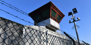 Ein Überwachungsturm auf einer Mauer, davor Maschen- und Stacheldraht, Kameras und Flutleuchten