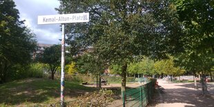 Im Vordergrund ein Schild mit der Aufschrift "Kemal-Altun-Platz", im Hintergrund ein Park mit grünen Bäumen und einem Schotterweg