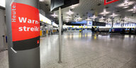 Eine weitgehend verwaiste Abflughalle eines Flughafens, im Vordergrund ein Schild „Heute: Warnstreik!“