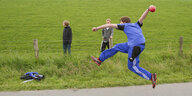Ein junger Mann in blauen Trainingsanzug wirft eine rote Kugel, seine Beine sind in der Luft