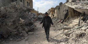 Ein Mann geht einen Weg zwischen eigenstürzten Gebäuden entlang, er breitet fassungslos die Arme aus. Das Bild stammt aus Jindaris, in der Region Aleppo