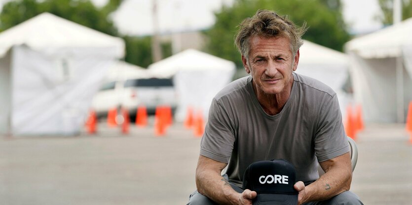 Sean Penn sitzt auf einem Stuhl im Freien, hinter ihm weiße Zelte