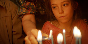 Ein Mädchen blickt ins Leere, vor ihr Kerzen. Eine davon zündet eine Person an, die hinter dem Mädchen steht