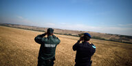 Zwei Männer in unterschiedlichen Uniformen der „European Border and Coast Guard Agency“ stehen auf einem Feld und blicken mit Ferngläsern in die Entfernung.