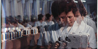Eine Frau in einem weißen Kittel arbeitet in einer Farbrik und setzt ein elektronisches Gerät zusammen, das Bild scheint wie durch Glas aufgenommen, wodurch sich der Bildinhalt dreimal wiederholt und visuell überlappt