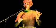 Die Klangkünstlerin Ute Wassermann steht auf einer Bühne und singt in ein Mikrofon. Sie hält eine Vogelpfeife in der Hand. Vor ihr steht ein Tisch, auf dem sich ein quadratischer, transparenter Behälter mit Wasser befindet. Wassermann hält ein Kontaktmikrofon über der Wasseroberfläche