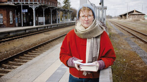 Eine Frau in roter Jacke steht auf einem Bahngleis mit einem Buch in der Hand