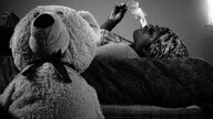 Eine schwarze Frau liegt rauchend auf dem Bett, neben ihr ein Teddy