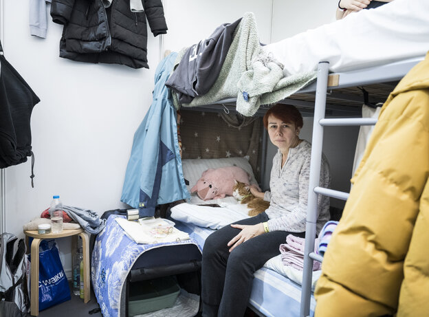 Eine Frau sitzt auf der unteren Matraze eines Doppelstockbetts. An dem Bett hängen Klamotten und Handtücher.
