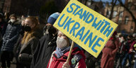 Eine Frau steht auf einer Demonstration mit einem großen Schild: Stand with Ukraine