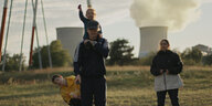 Ein Mann mit Kind auf den Schultern, ein Junge und eine Frau. Hinter ihnen ein Atomkraftwerk im Abendlicht.