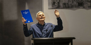 Der Schriftsteller Rainald Goetz hält an einem Lesepult während eines Vortrags ein Buch hoch