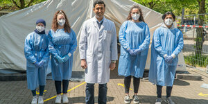 Wjahat Waraich steht im Arztkittel zusammen mit mehreren Helferinnen in Schutzkleidung vor dem Zelt seines improvisierten Testzentrums auf einem Parkplatz