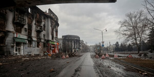 Eine menschenleere Straße, links und hinten sind zerstörte Gebäude zu sehen, kahle Bäume rechts, düsterer Himmel