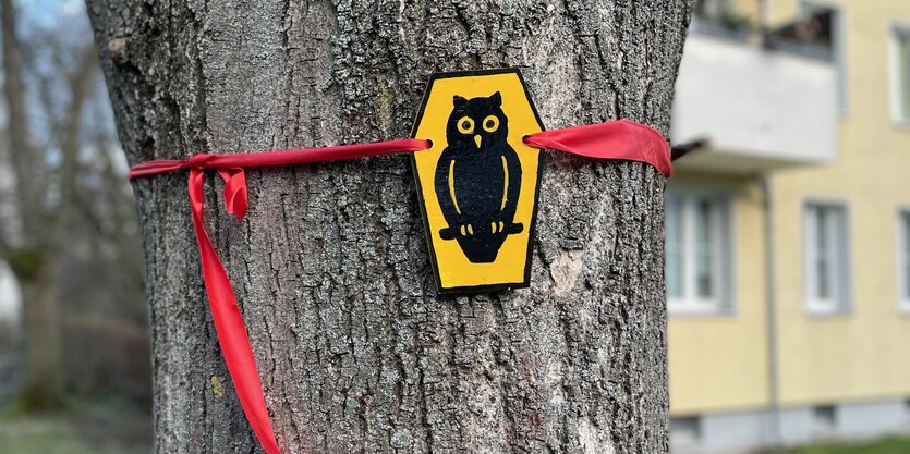 An einem Baum ist mit rotem Band eine schwarze Eule auf gelbem Grund befestigt