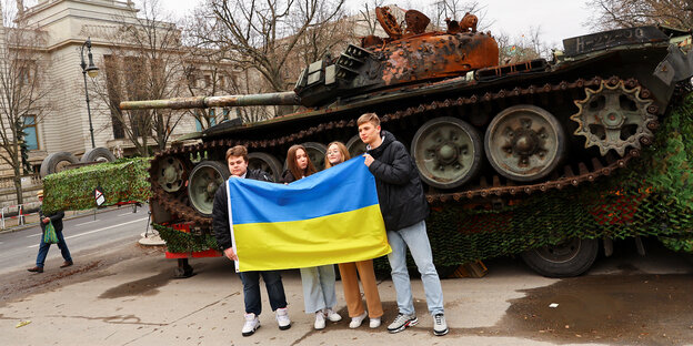 Vier junge Leute halten sich die ukrainische Flagge vor den Körper und posieren vor einem zerstörten russischen Panzer, der vor der russischen Botschaft in Berlin steht