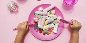 Ein Kind - nur die Hände sind zu sehen sitzt vor einem pinkfarbenen Teller auf dem bunte Marshmallows und Jelly Beans liegen